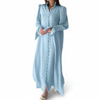 Woman wearing a light blue dress with long sleeves against a white paneled wall.