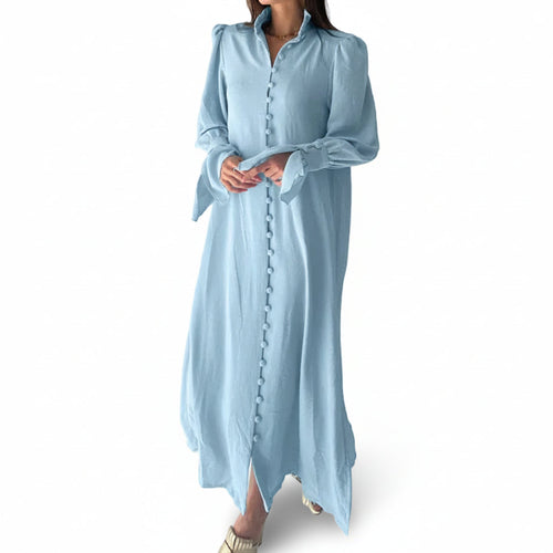 Woman wearing a light blue dress with long sleeves against a white paneled wall.