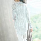 Person wearing a light blue floral lace blouse with white pants, standing in a softly blurred indoor setting.